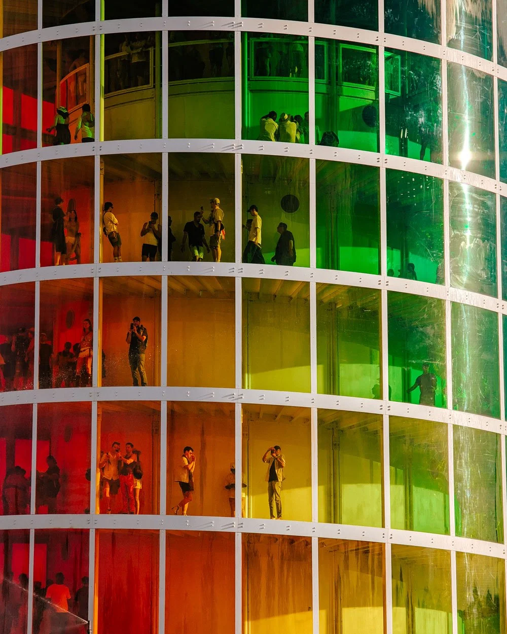 People inside a cylindrical building with colorful panes of glass in red, orange, yellow, and green, looking around, taking photos, and talking.