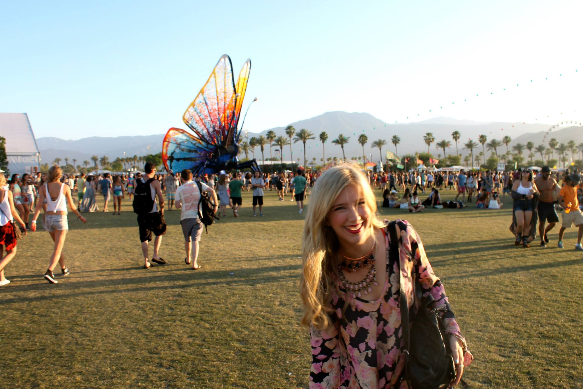 A woman with blonde hair and a floral dress smiling at a music festival with numerous people, a giant butterfly sculpture, tents, and palm trees in the background.