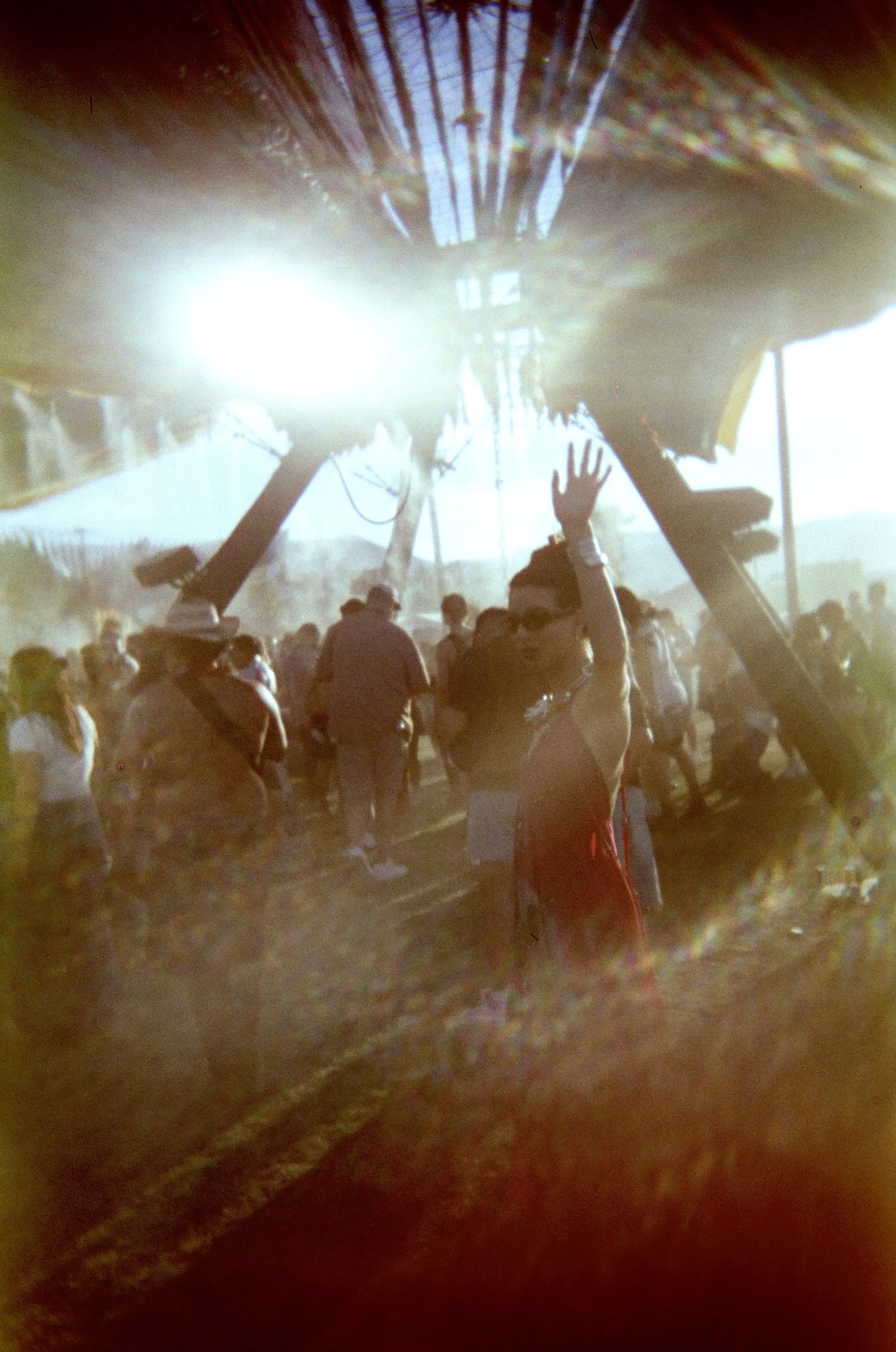 A woman in a red dress with sunglasses waving at an outdoor event as a crowd stands beneath a canopy supported by large beams, with bright sunlight creating a hazy atmosphere.