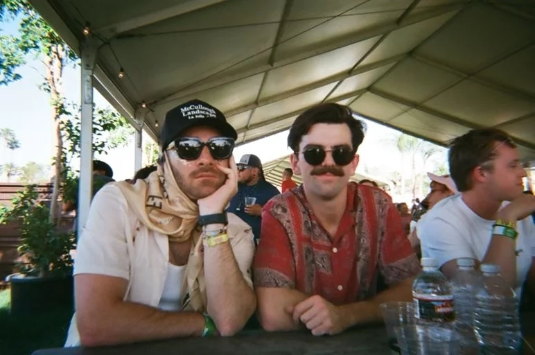 Two men sit at a table outdoors under a tent during daytime. They wear sunglasses and casual clothing; one has a cap and a scarf, the other a mustache and patterned shirt. Several people are in the background.