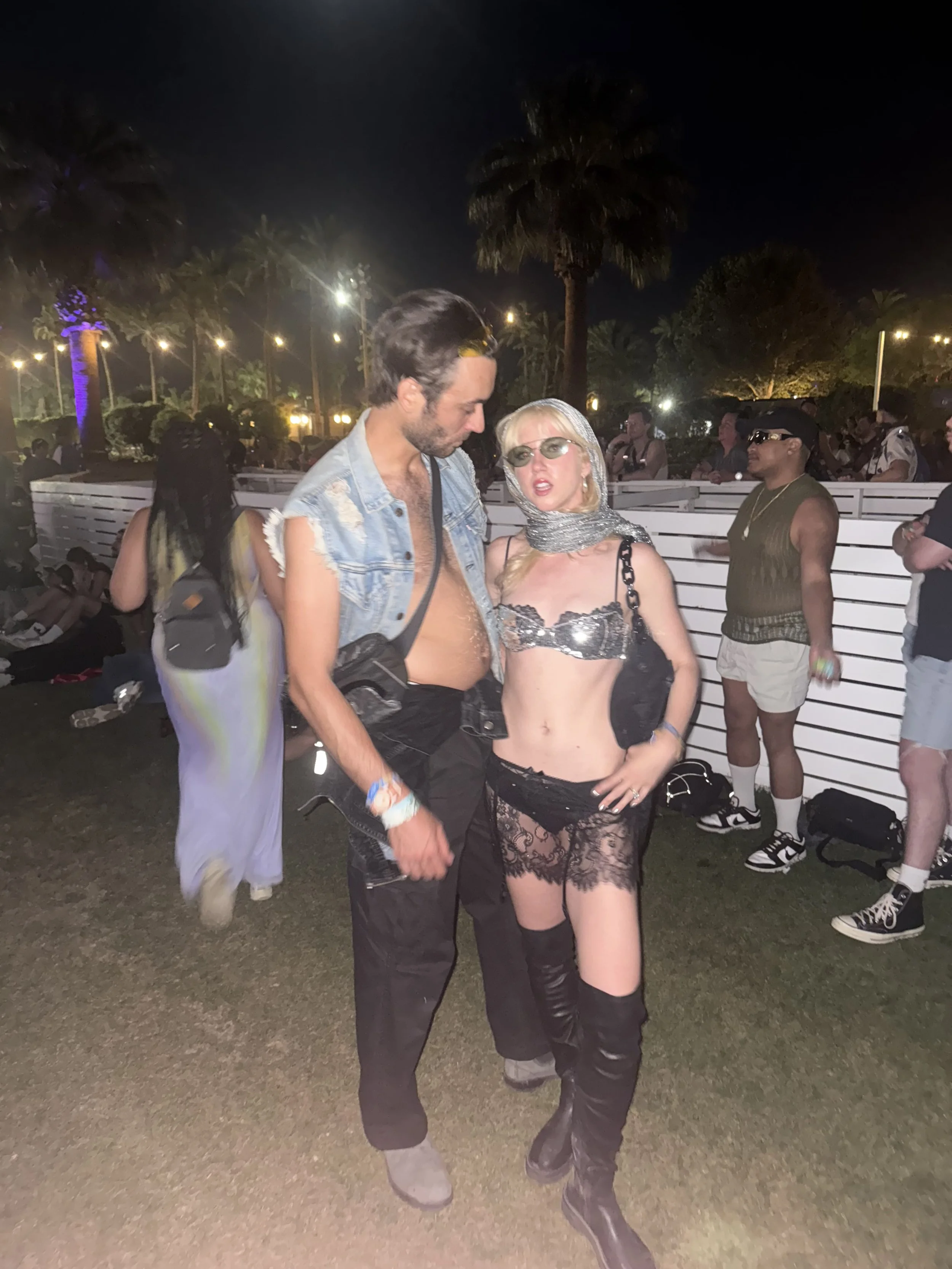 Two people standing outdoors at night, dressed in festival or party attire, with palm trees and other people in the background.