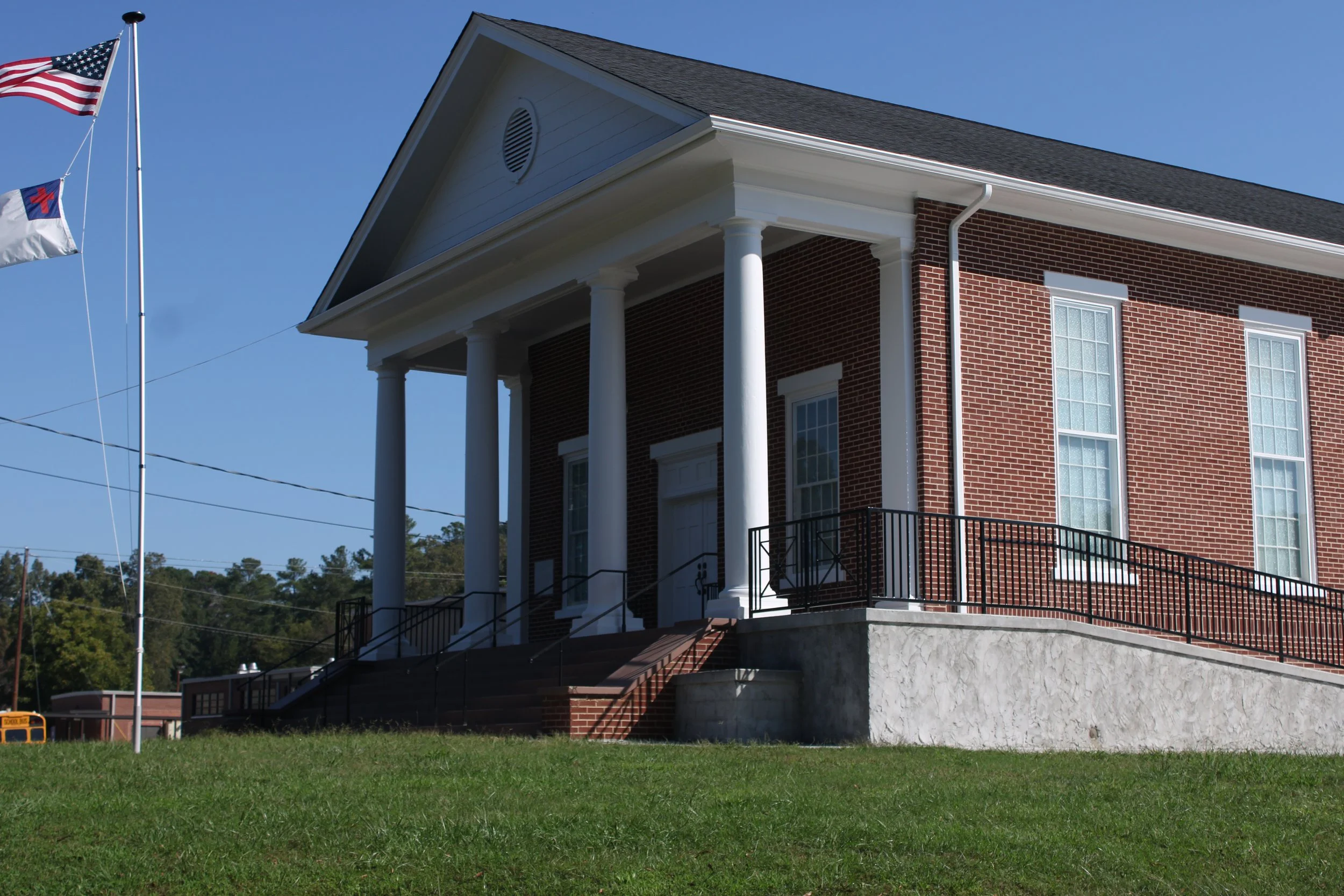A brick building with a white pediment and columns, four tall windows, and stairs leading up to the entrance. An American flag and another flag are flying on flagpoles nearby.
