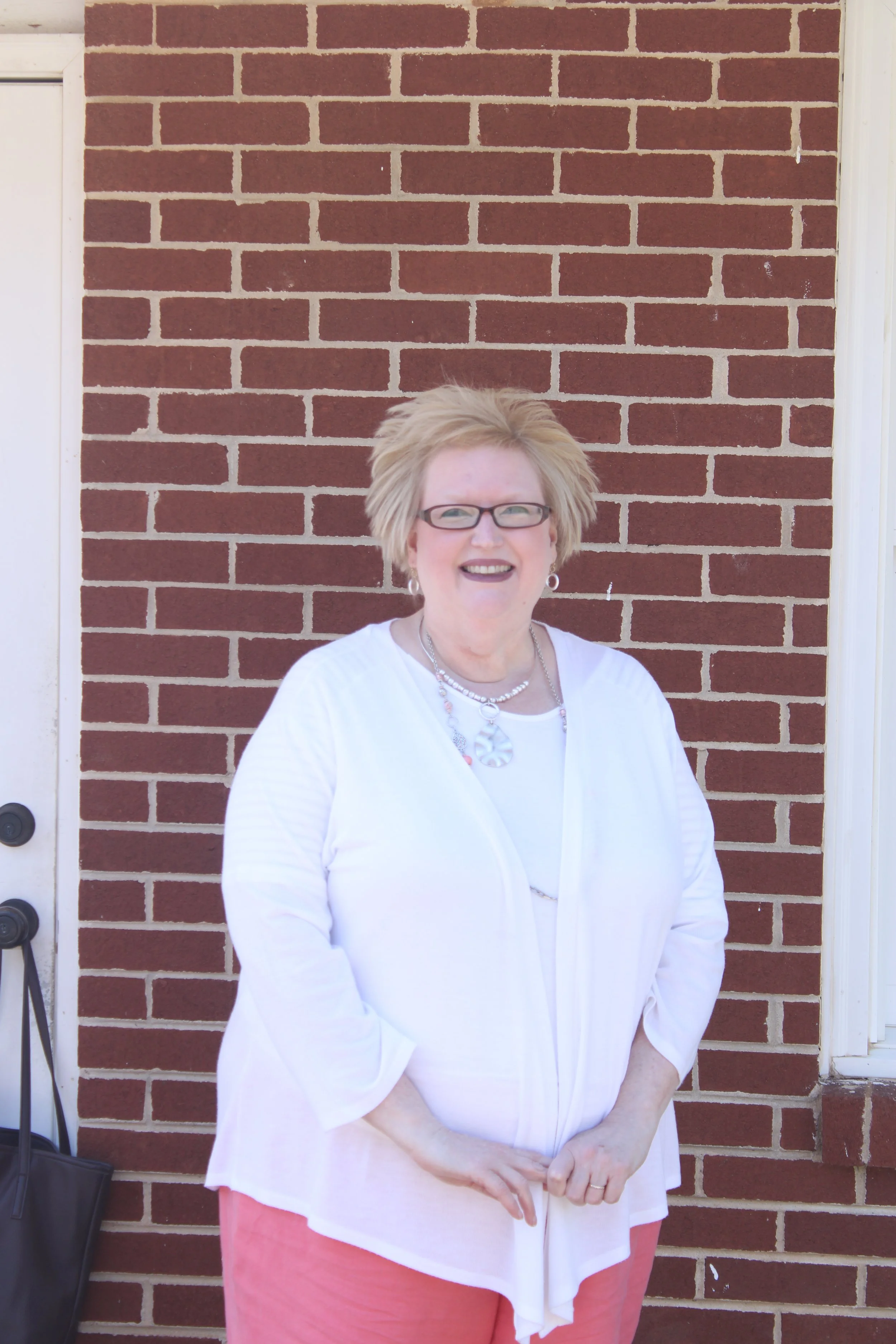 A smiling woman with glasses and short blonde hair standing in front of a red brick wall.