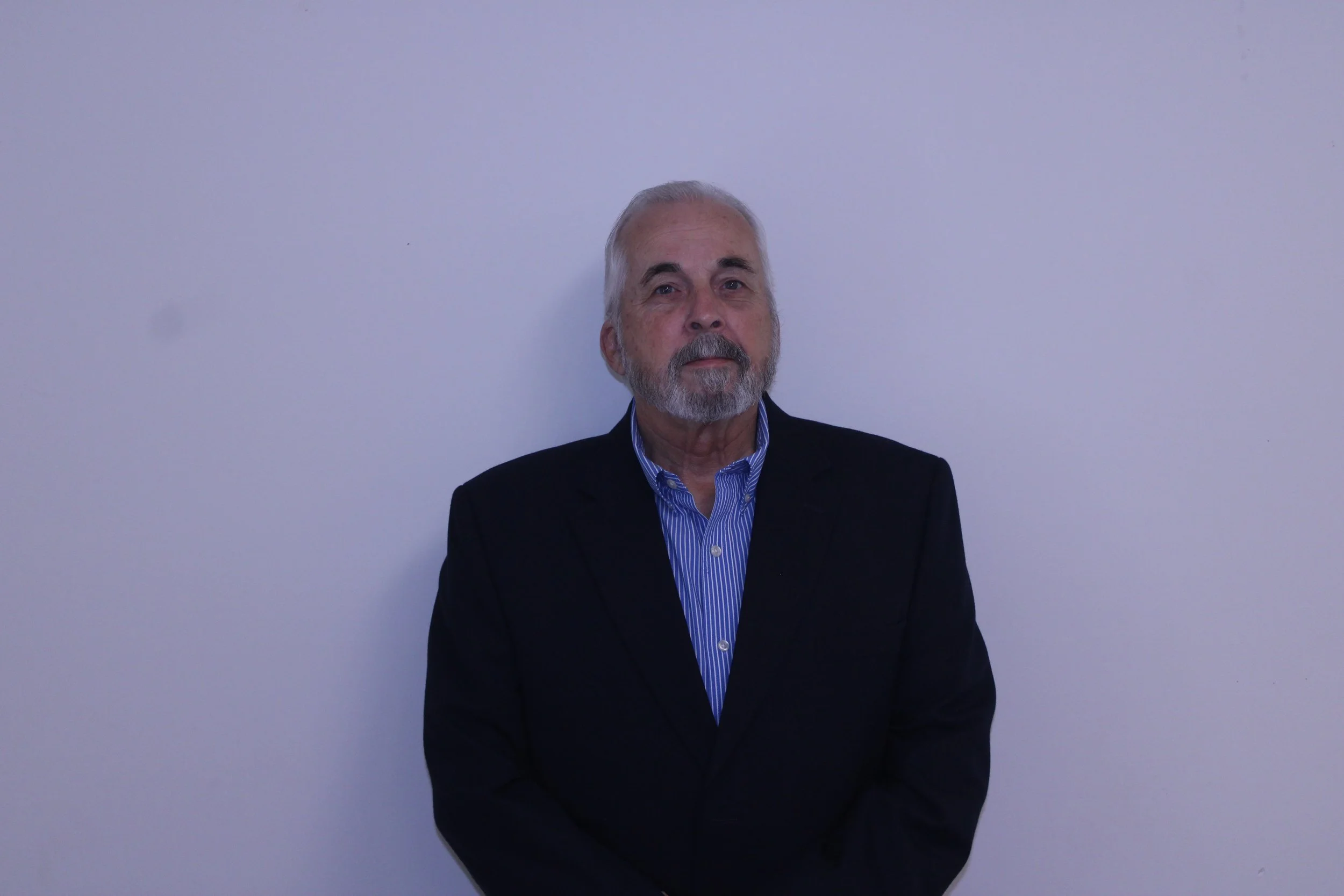 An older man with gray hair and a gray beard, wearing a dark suit jacket and a blue striped shirt, stands against a plain light-colored wall.