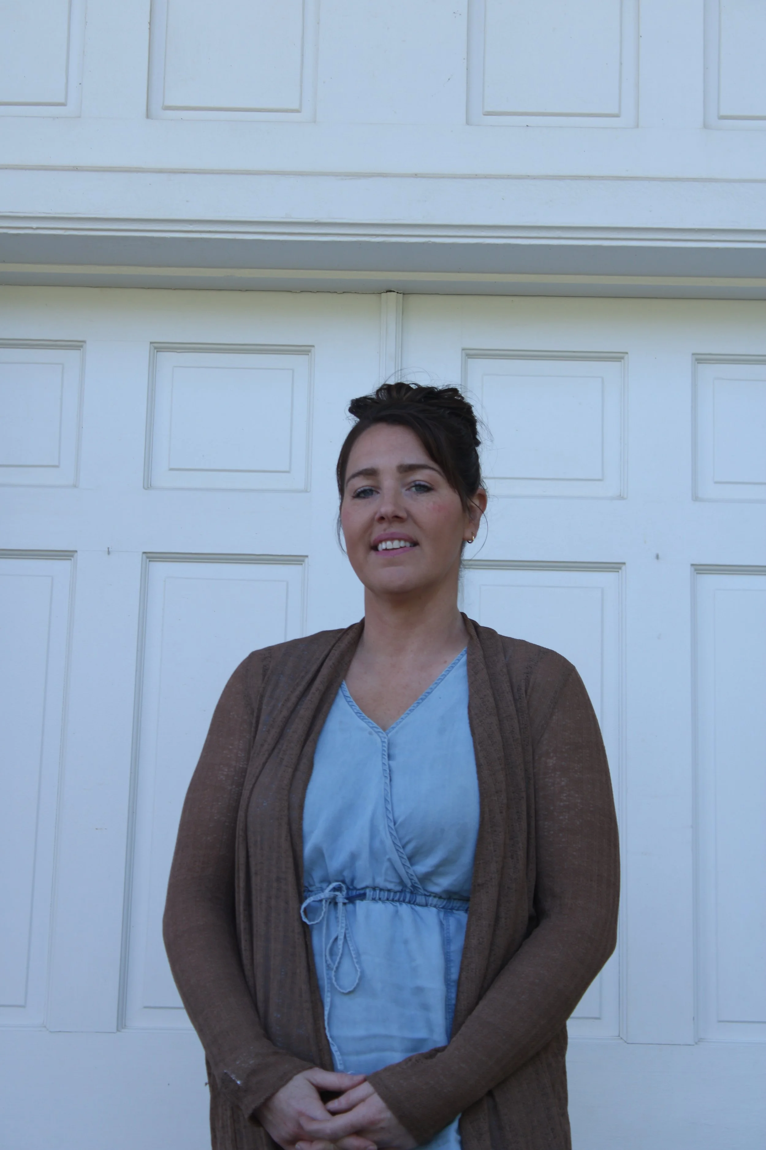 A woman standing in front of a white garage door, wearing a brown cardigan over a blue dress, with her hands clasped in front of her.