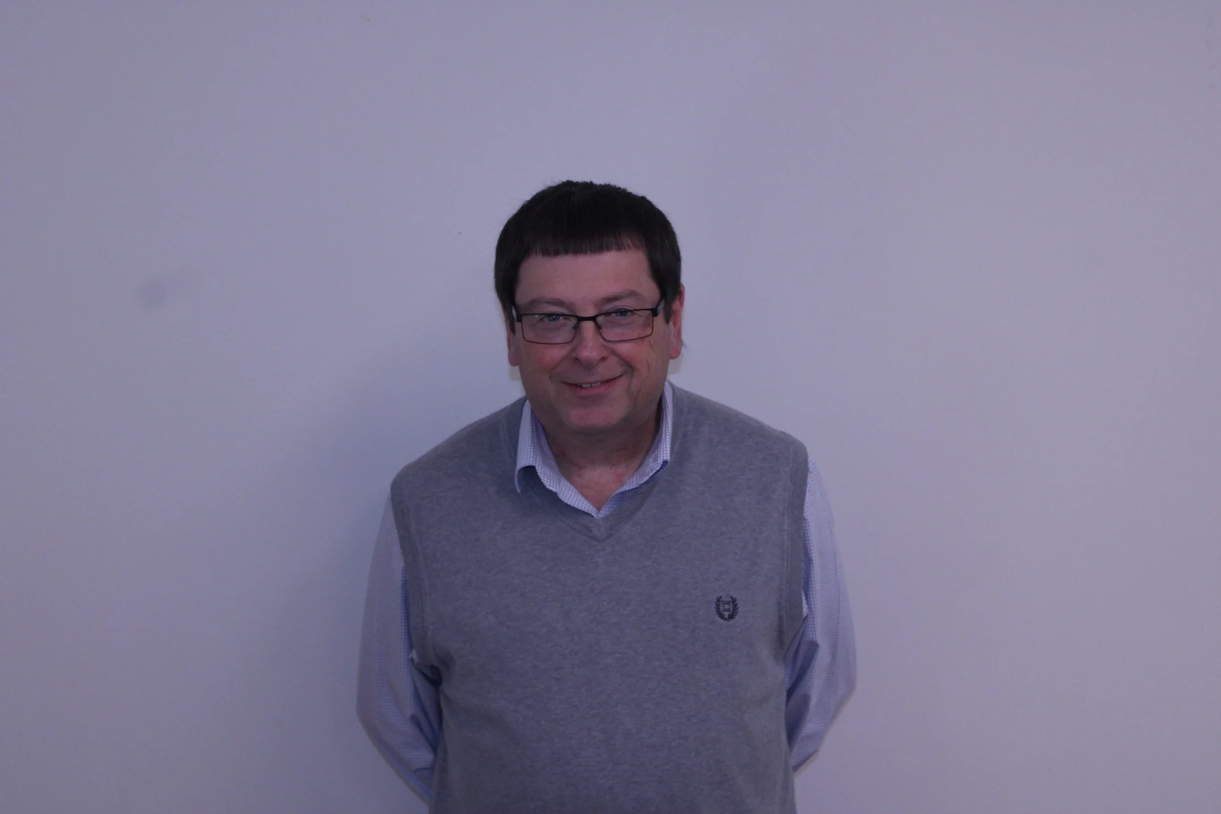 A man with glasses, wearing a gray sweater vest over a collared shirt, standing against a plain white wall and smiling.