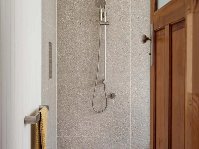Shower with gray tiled wall, handheld shower head, door slightly ajar, and towel hanging on a hook.