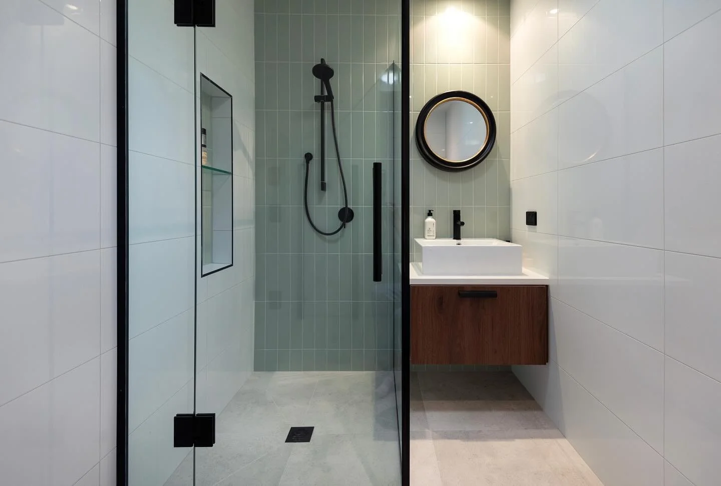 Modern bathroom with a walk-in shower with black fixtures and a glass door, a round mirror above a white sink with a wooden cabinet, and white tiled walls.