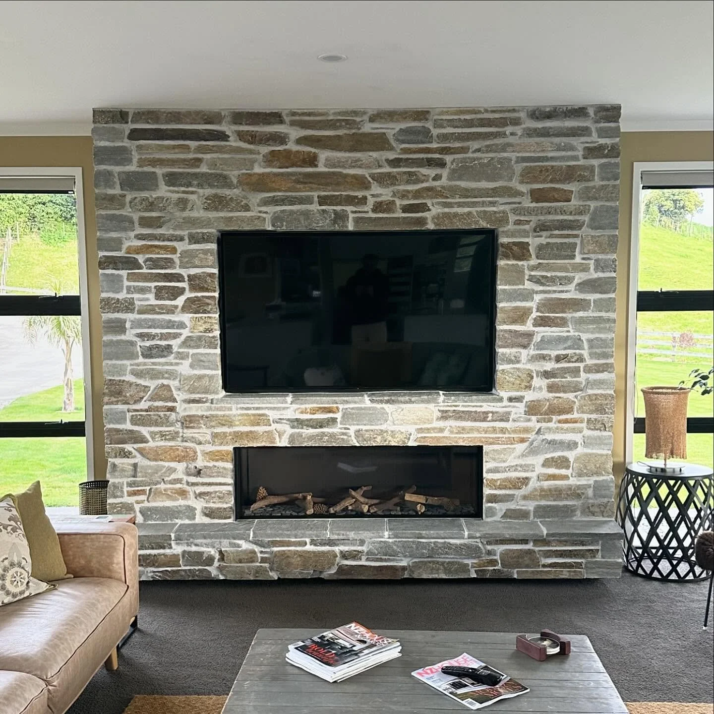 Living room with a gas stone fireplace, mounted flat-screen TV, large windows showing green outdoor scenery, a light pink sofa, a coffee table with magazines, and side tables with decor.