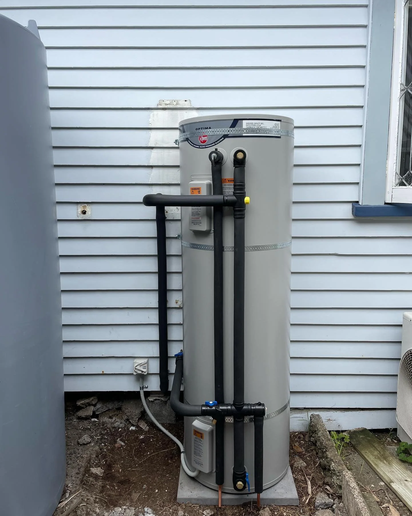 A vertical hot water heater installed outside a house with white siding, connected to black pipes and a gas heater.