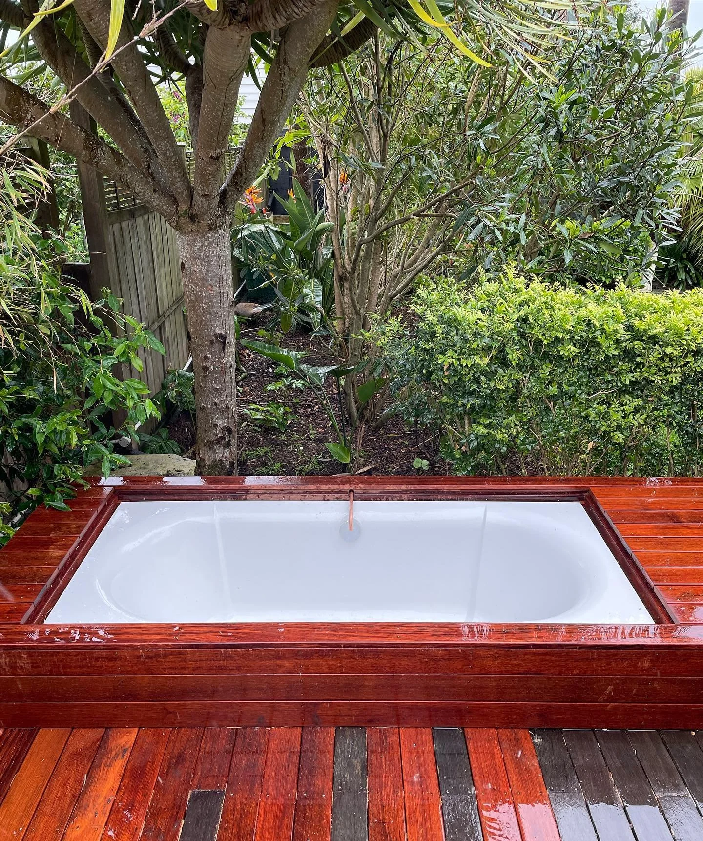 Outdoor hot tub surrounded by a wooden deck and lush greenery in a garden setting.