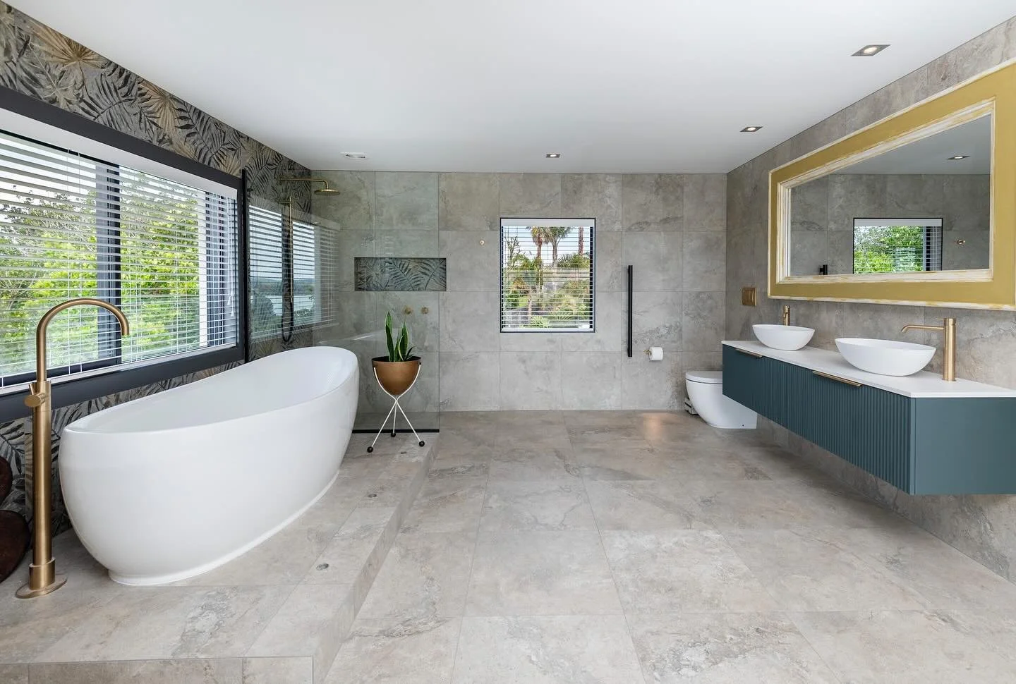 Modern bathroom with large windows, a free-standing bathtub, a dual sink vanity with vessel sinks, a toilet, and minimal decor, featuring neutral tile flooring and walls.