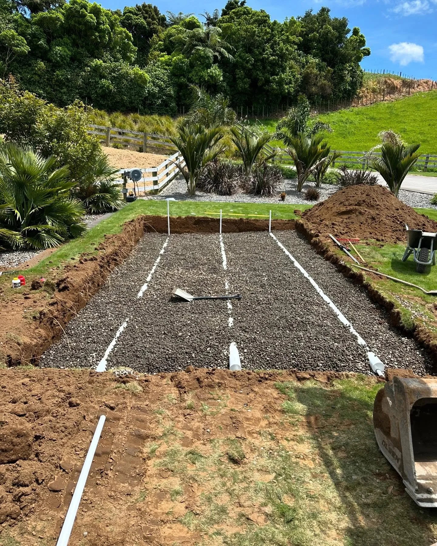 A garden with palm trees, a fenced hillside, and a small construction area for a concrete slab with white lines, pipes, and construction tools.