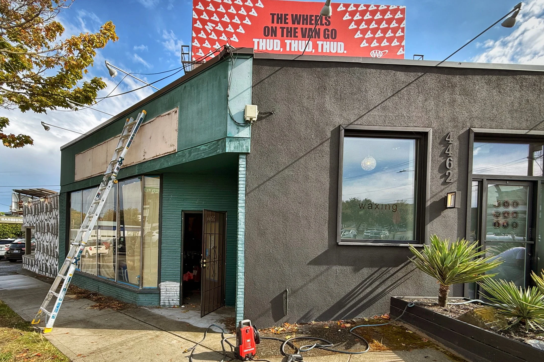 Exterior of a tattoo salon with large windows and a door, some plants outside, a ladder leaning against the building, and equipment on the sidewalk, with a large billboard on top advertising a VW van and music, under a partly cloudy sky.