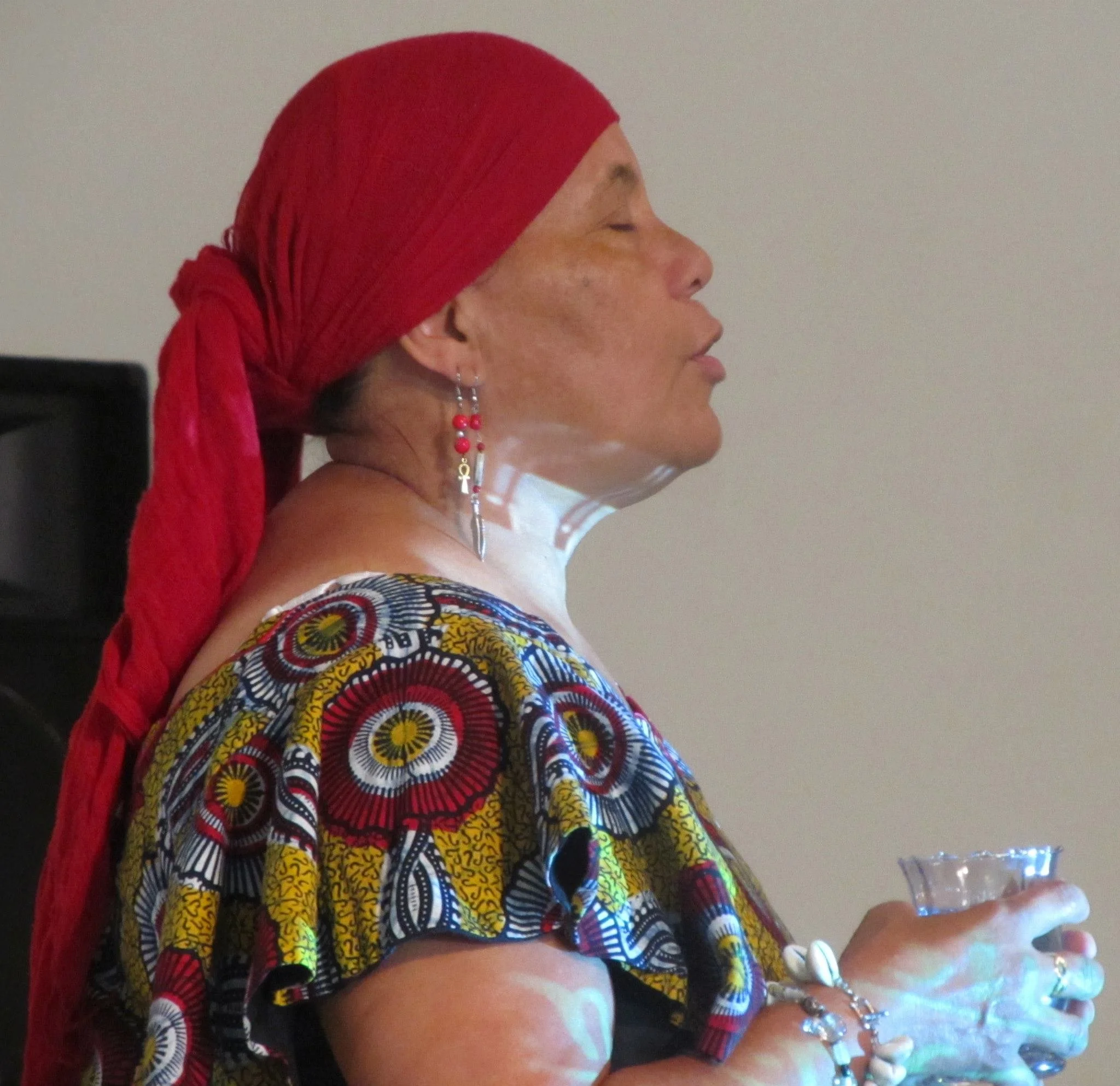 A woman wearing a red headwrap and colorful, patterned top holding a glass.