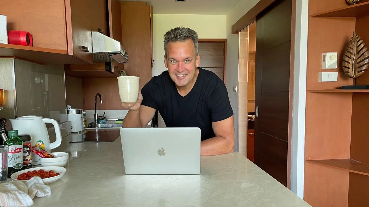 A man (Gregg Zambrovitz) with short gray hair wearing a black T-shirt smiling at the camera while sitting at a kitchen counter with a laptop, holding a ceramic coffee mug.