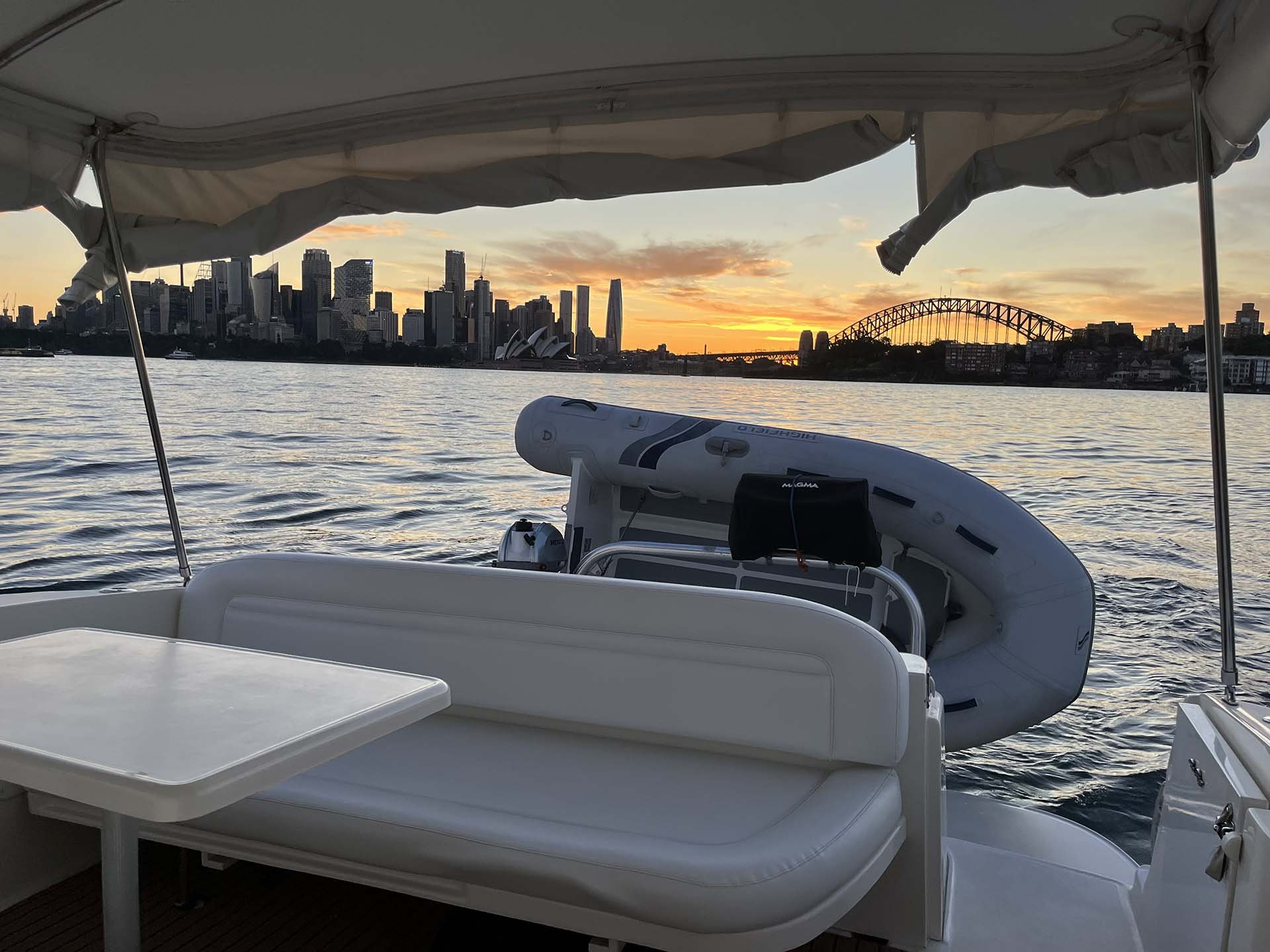 View from a Opal showing a city skyline at sunset, with buildings and the Sydney Harbour Bridge in the background, and part of the boat's seating.
