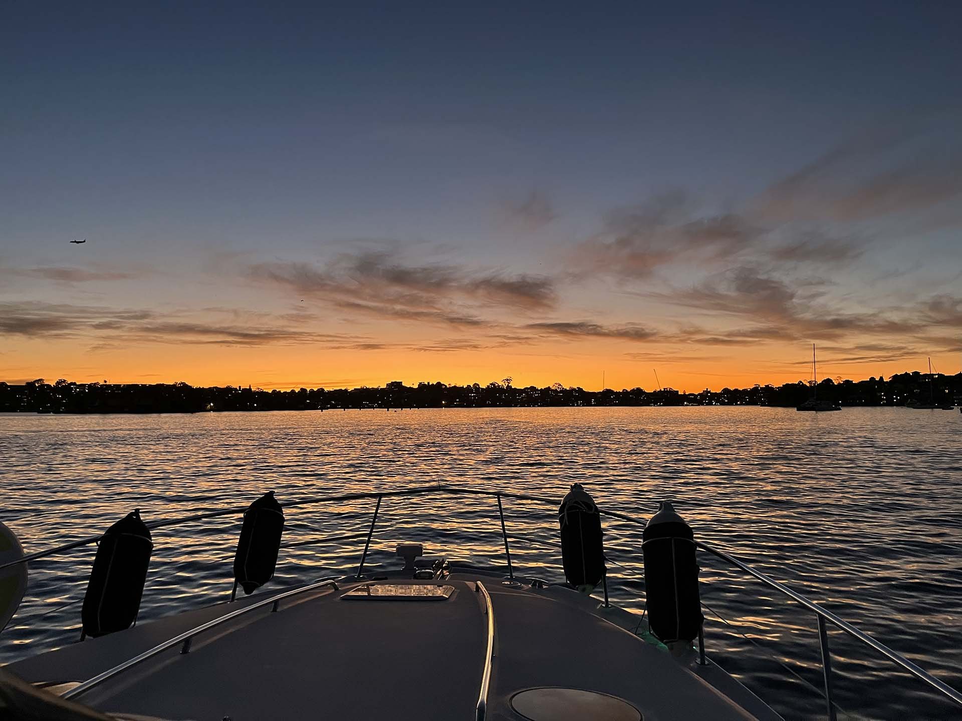 View from a boat at sunset with orange, pink, and purple sky. Calm water reflecting the sky. Silhouettes of distant boats and trees on the horizon. A small airplane flying in the distance.