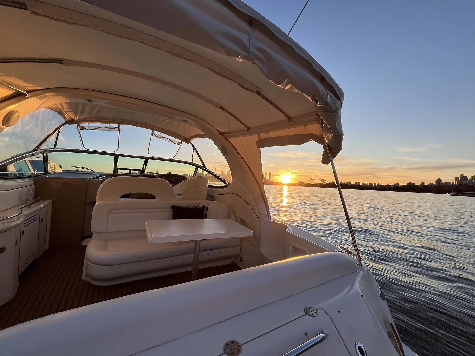 View from Opal at sunset showing the boat's interior with white seating, a small table, and a dashboard, overlooking a waterway with a city skyline and a harbour bridge in the distance.