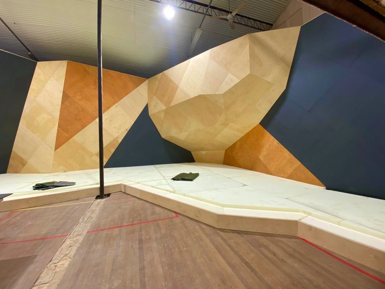 Indoor climbing gym with a geometric climbing wall, featuring various angled holds and an overhanging section, with black climbing grips on the left side and black clothing or mats resting on the padded floor. Boulderlounge Gympie Queensland