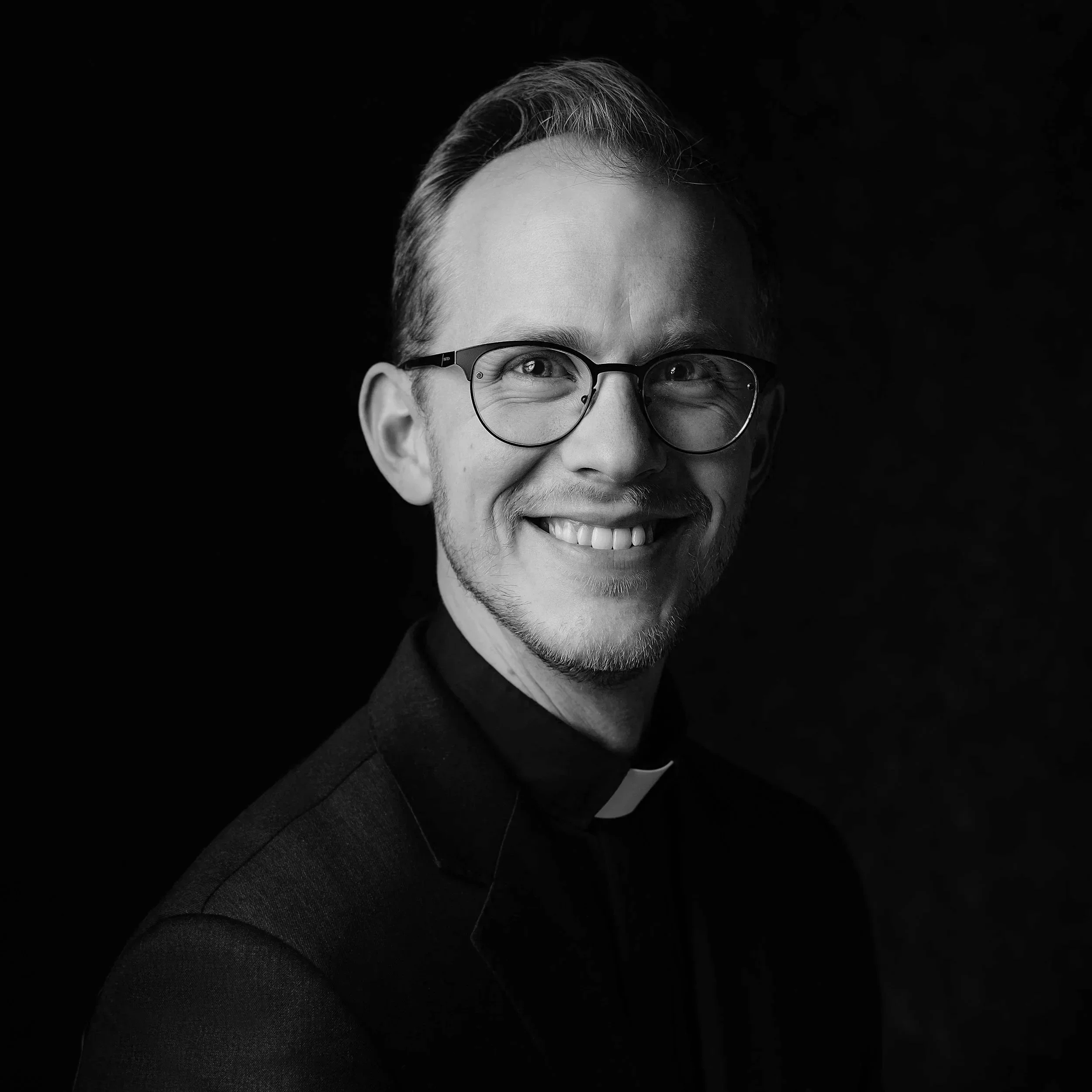 Black and white portrait of a smiling man with glasses, wearing a suit, against a dark background.