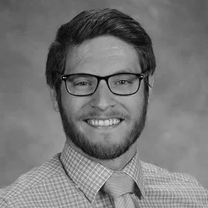 Black and white photograph of a smiling young man with glasses and a checkered shirt.