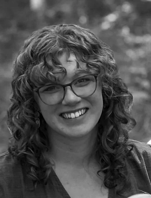 Black and white photo of a woman with curly hair and glasses smiling outdoors.
