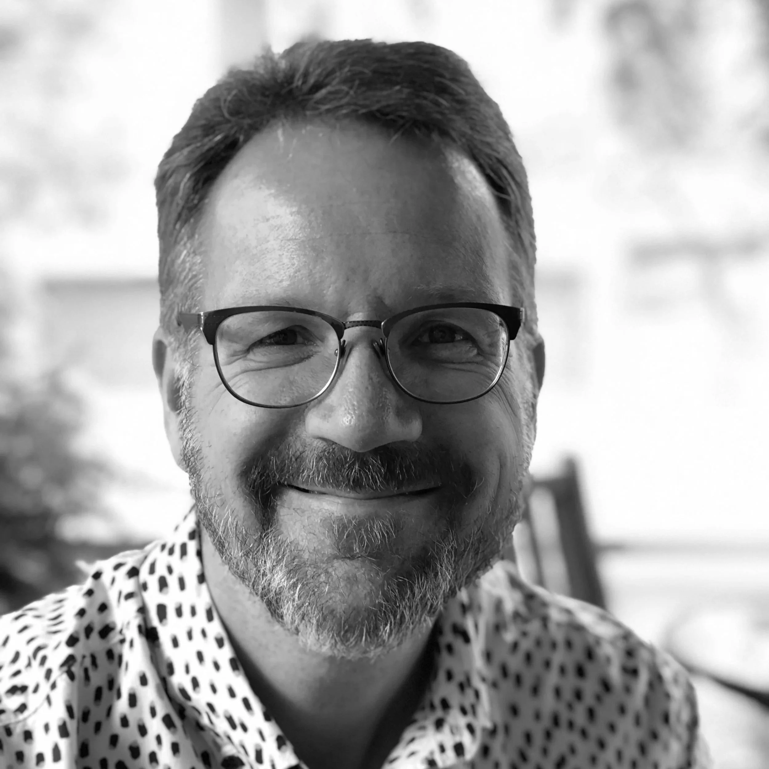 Close-up black and white photo of a middle-aged man with glasses, gray hair, and a beard, smiling and wearing a polka dot shirt.