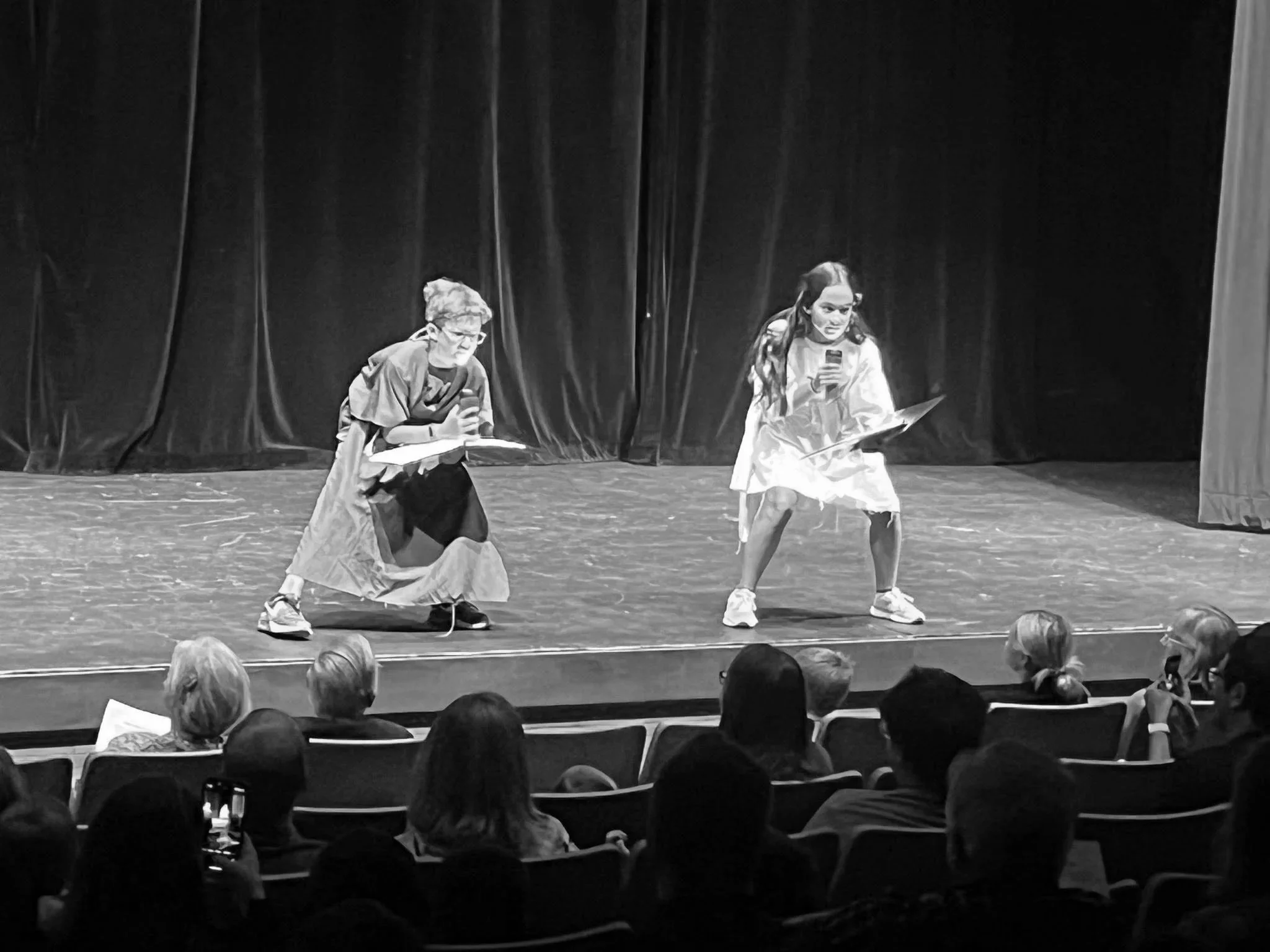 Two children perform on stage, one kneeling with a paper and a microphone, the other standing with a paper and a microphone, with an audience watching.