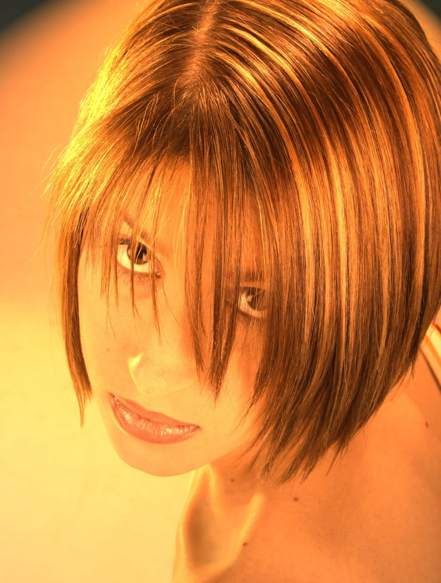 Close-up of a woman with a short, layered, reddish-brown hairstyle looking at the camera with a slight smile against a warm, orange background.