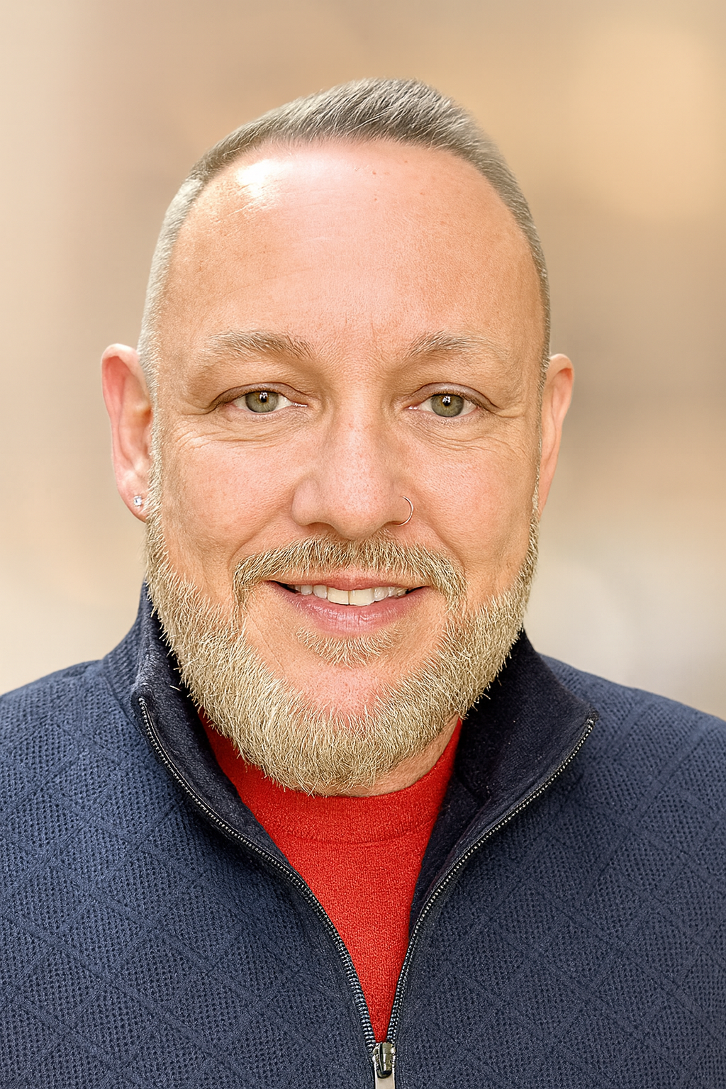 Close-up portrait of a smiling man with a beard, mustache, and piercing eyes, wearing a nose ring and a dark zipped sweater over a red shirt.