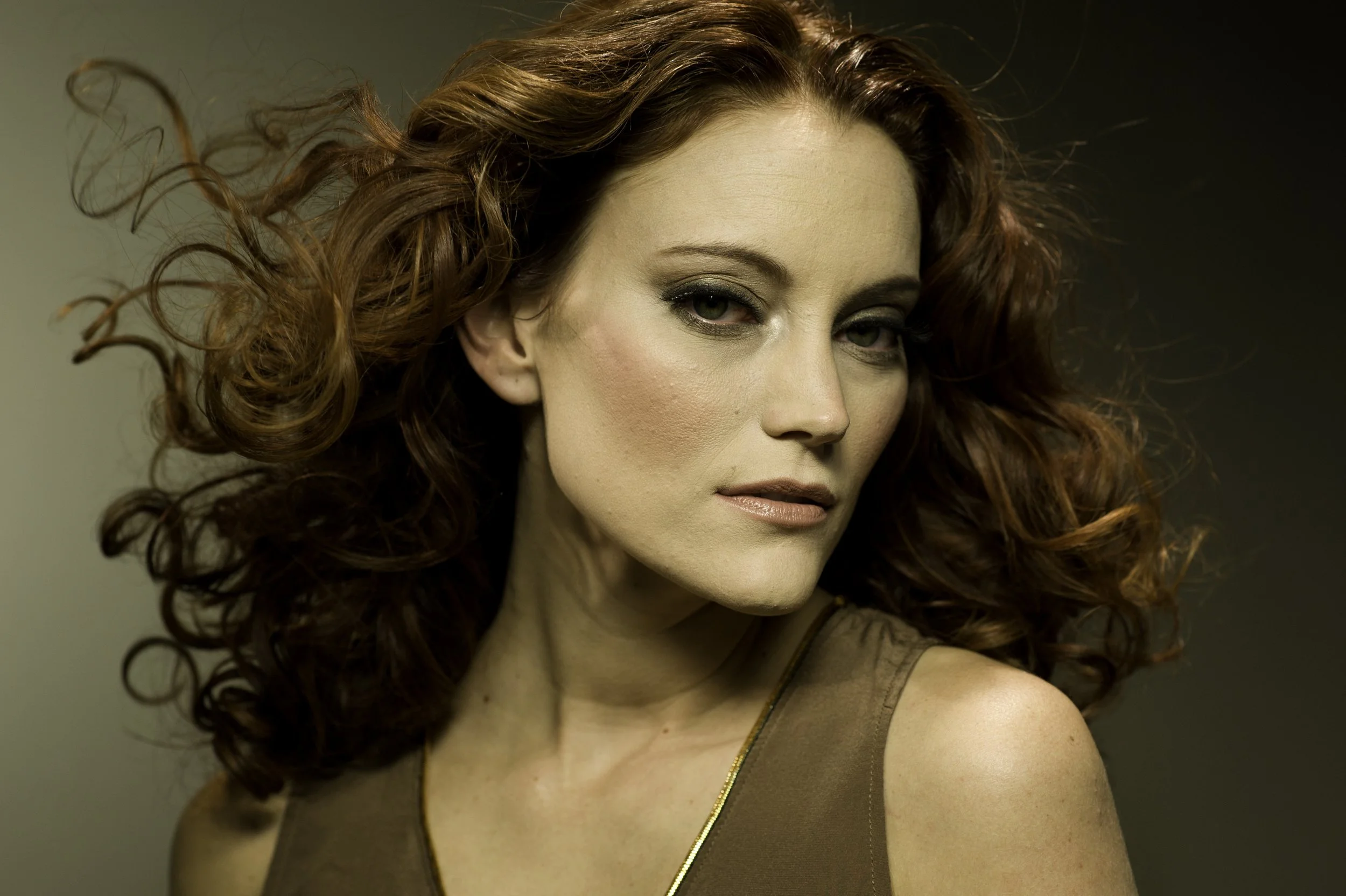 A woman with curly red hair and fair skin looks over her shoulder, wearing makeup and a sleeveless brown top.