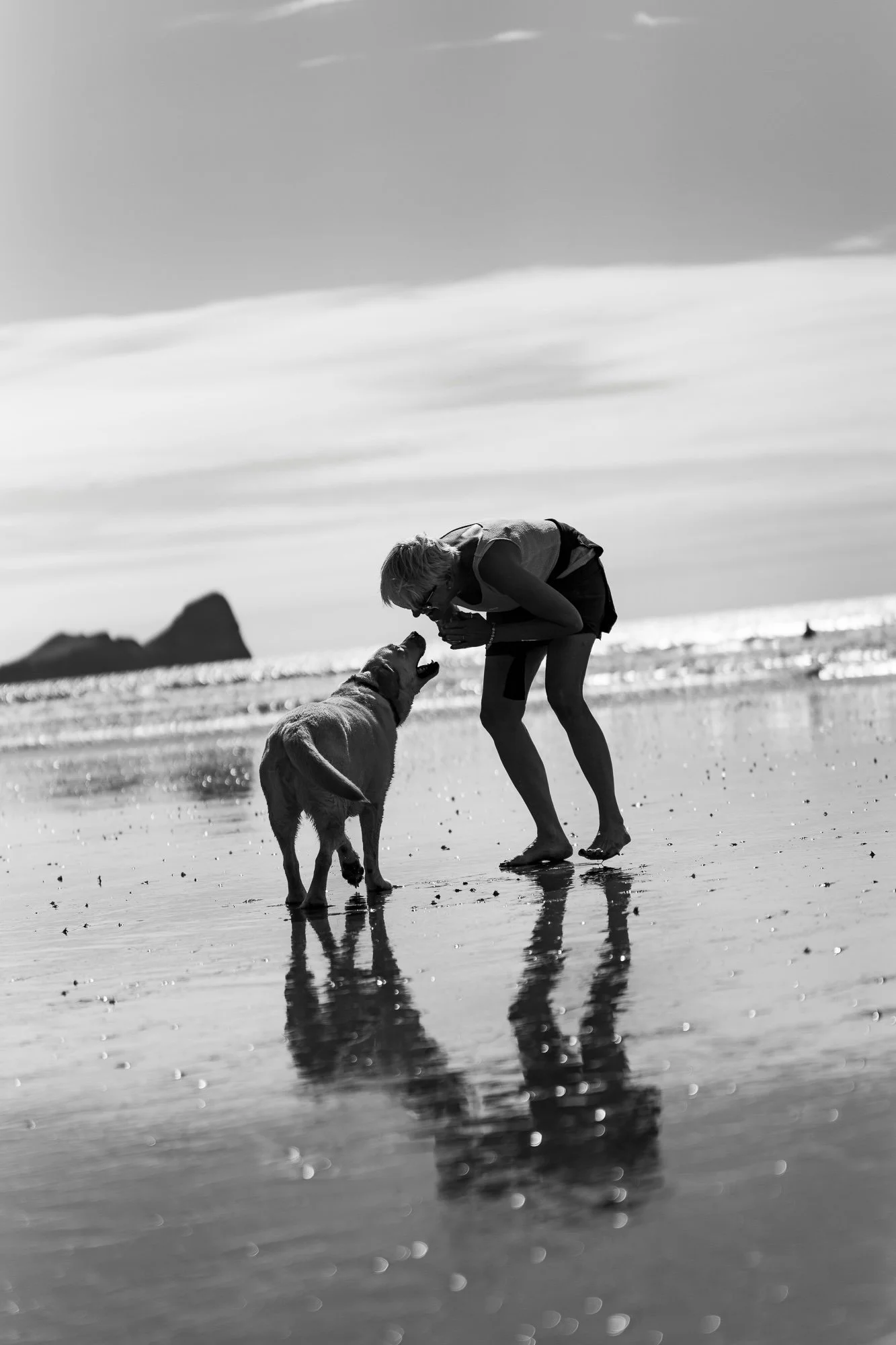 A woman and a dog playing on a beach, with water and a rocky island in the background, in black and white.