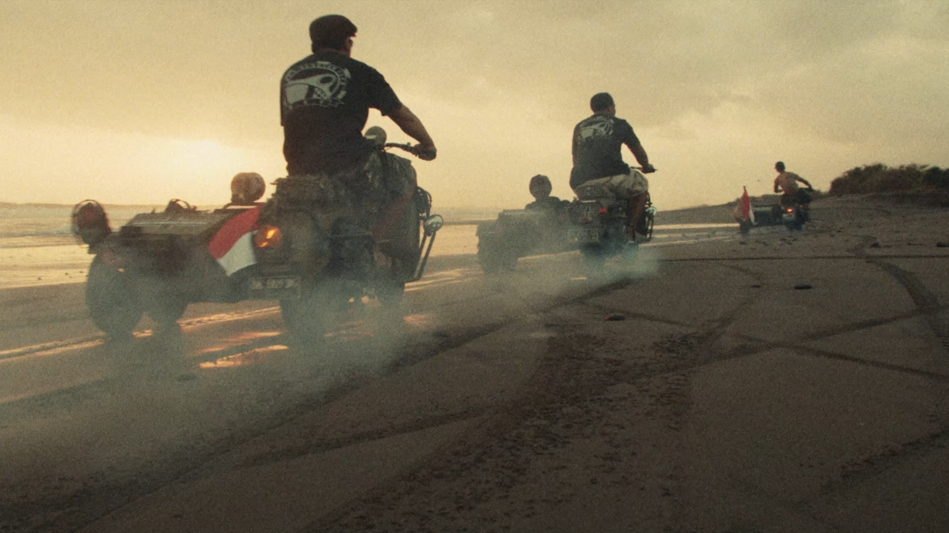 Four people riding vespas on a beach with tire tracks in the sand and the indonesian ocean in the background.