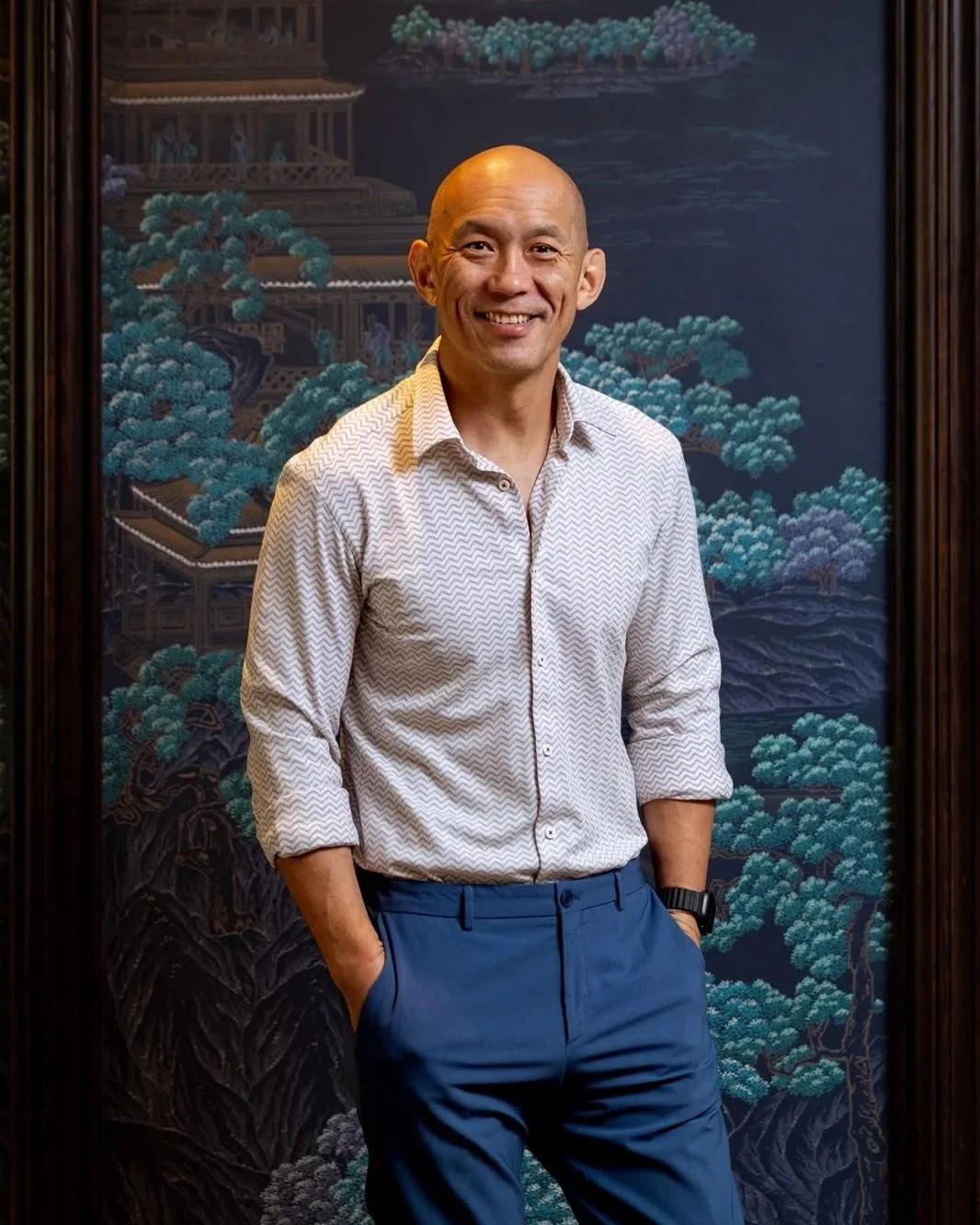 A man smiling, wearing a light-colored patterned dress shirt and blue pants, standing in front of a dark, traditional Asian landscape painting with trees and buildings.