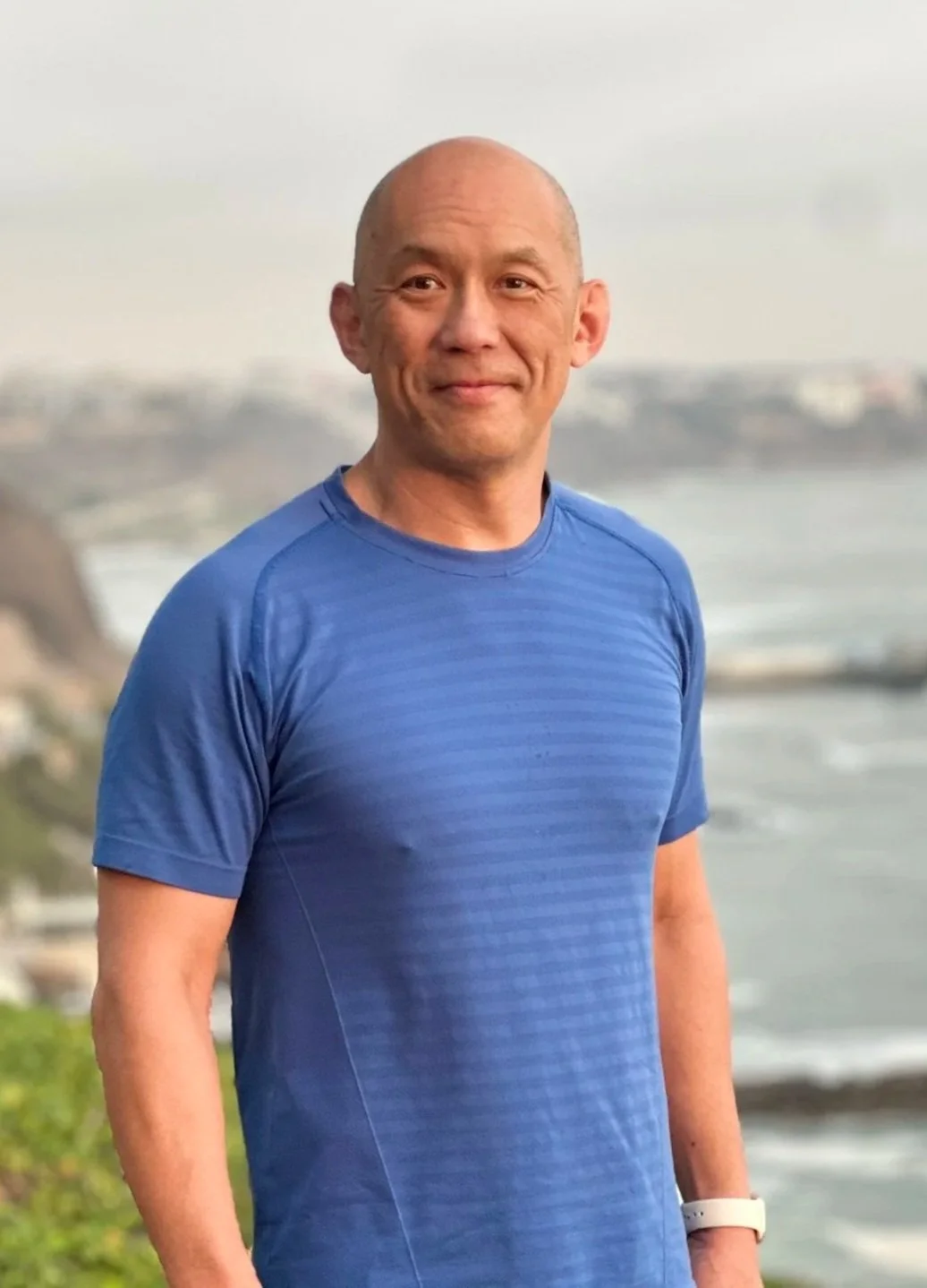 A middle-aged man with a shaved head smiling outdoors by the water, wearing a blue athletic shirt.