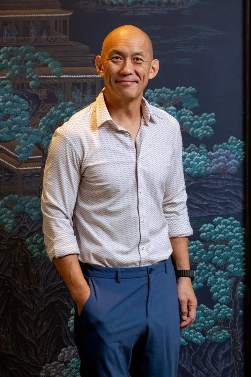 A bald man in a white patterned shirt and blue pants standing with one hand in his pocket, smiling, in front of a traditional Asian-themed painted backdrop featuring trees, mountains, and buildings.