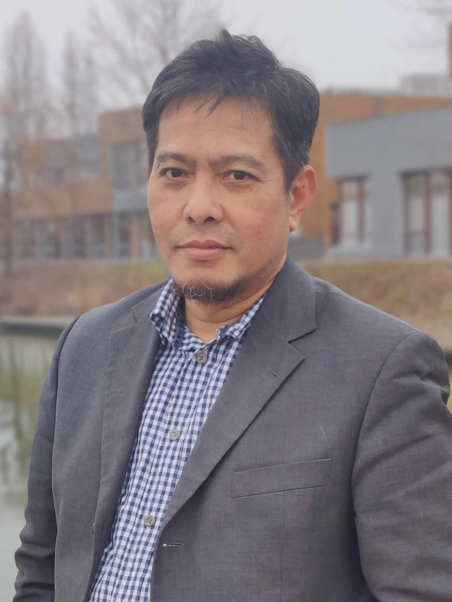A man in a gray blazer and checkered shirt standing outdoors near a body of water with modern buildings in the background.