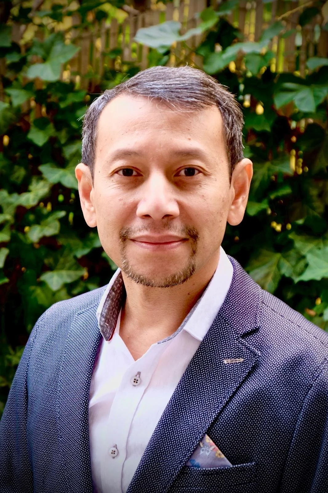A man with short dark hair, a slight beard, and a mustache standing outdoors in front of green leafy plants. He is wearing a navy blue suit jacket with a white shirt and a patterned pocket square, smiling at the camera. Chairman of Diastra.