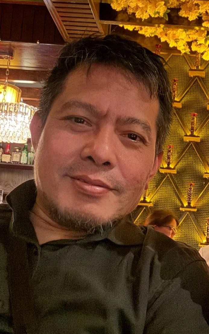 A man with short dark hair and a goatee, smiling, in a restaurant with a wooden ceiling, yellow flowers, decorative shelves, and bottles behind him. One of the founder of Diastra.