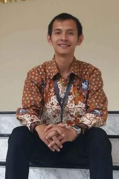 A young man sitting cross-legged on a bench, smiling at the camera, wearing a patterned batik shirt, black pants, and a lanyard around his neck. One of the founder of Diastra.