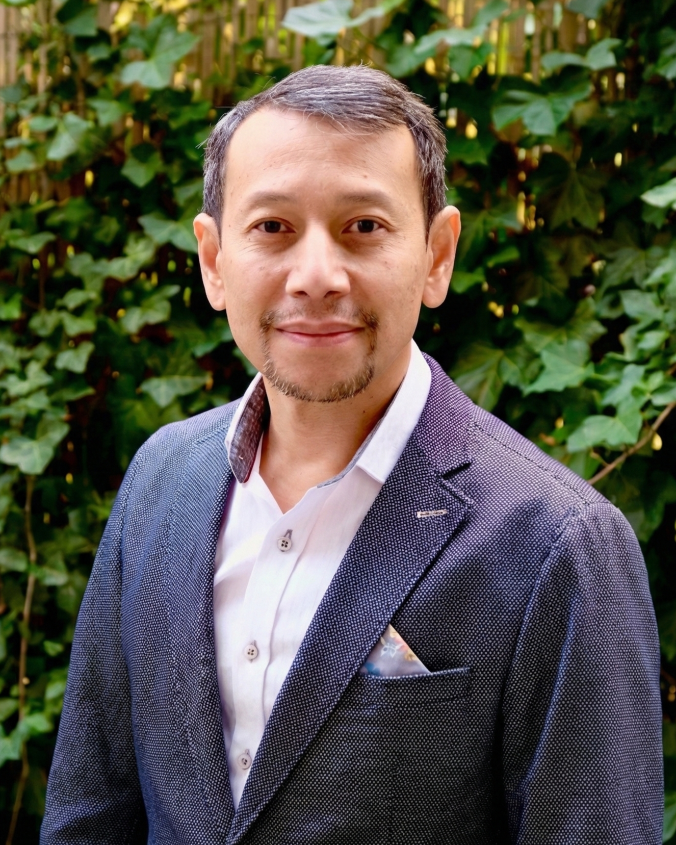 A man in a dark suit and white shirt standing outdoors in front of green foliage.