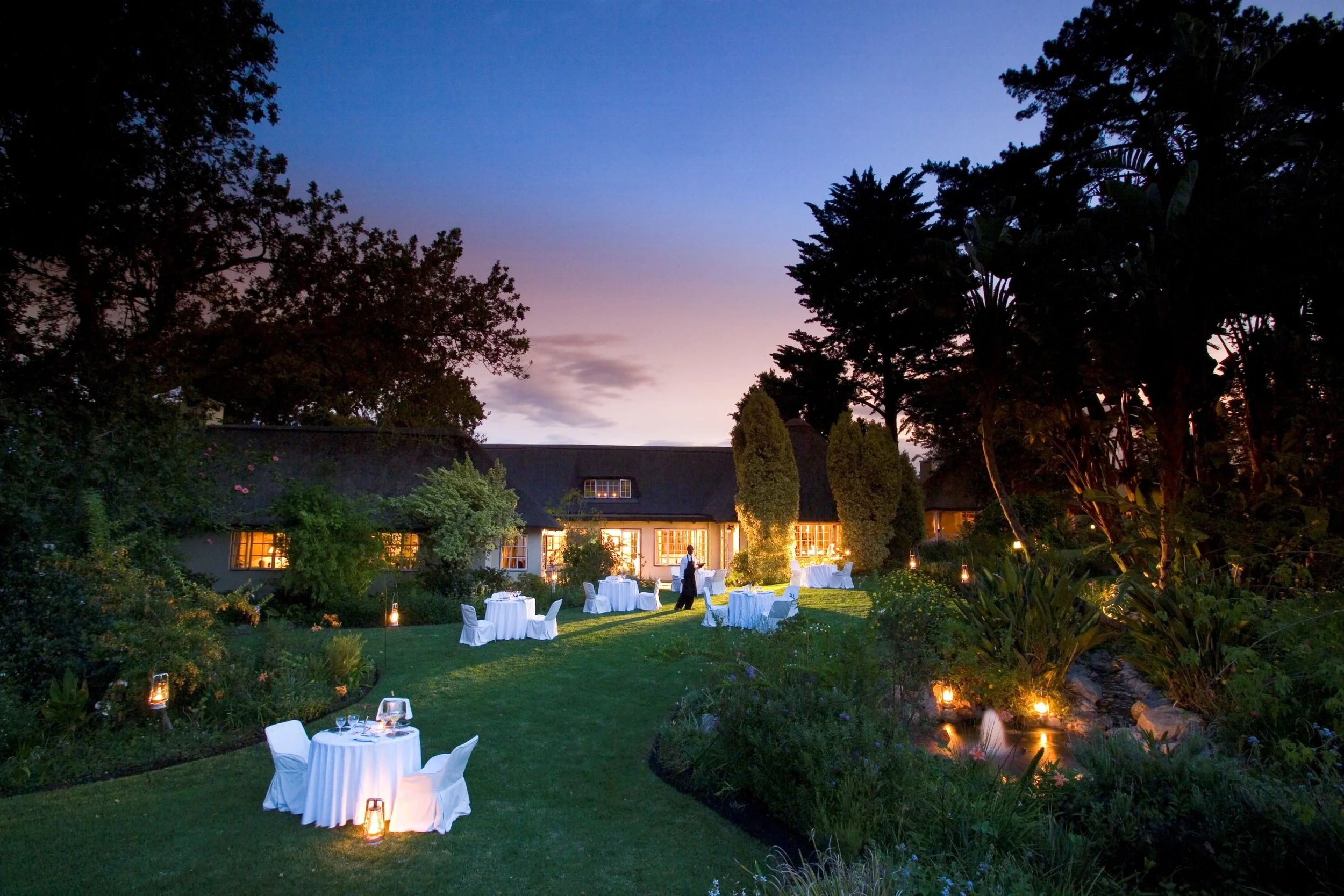 Outdoor evening dining area with tables and chairs covered in white cloth, lit by lanterns, in a lush garden with a house in the background and a colorful, dusky sky.