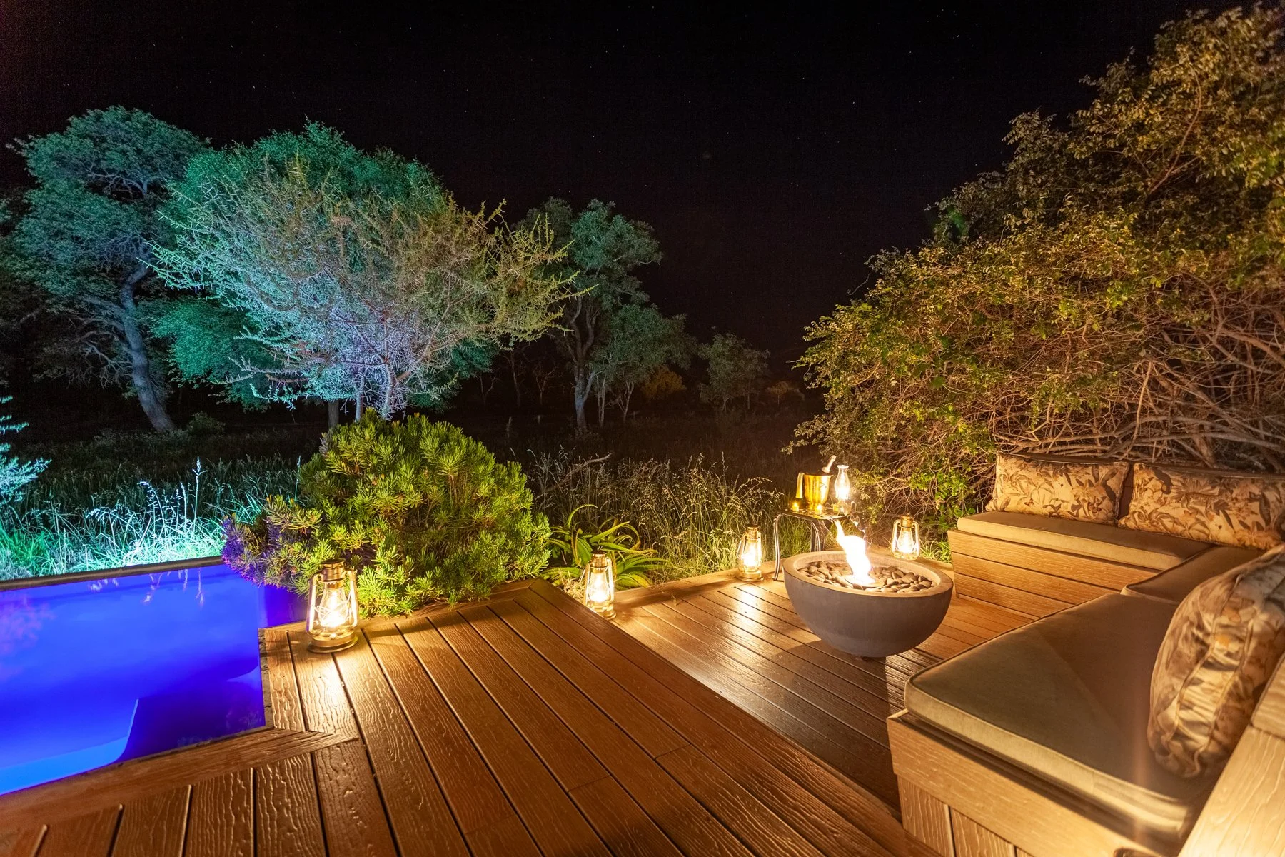 Nighttime outdoor patio with wooden deck, illuminated by lanterns, overlooking a dark wooded area with trees, a fire pit, and a blue lit hot tub.