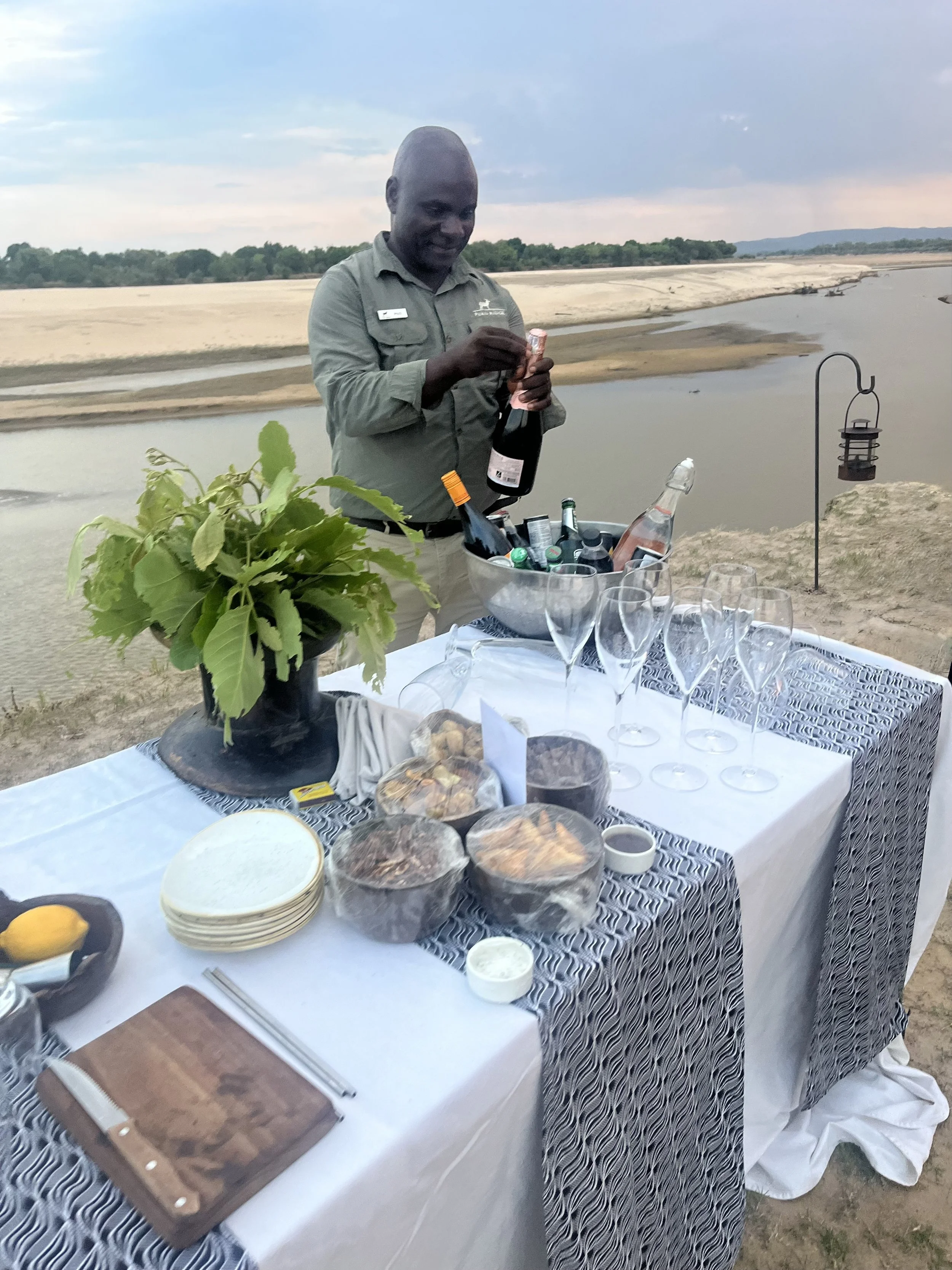 A man setting up a buffet table outdoors by a river, with food, drink, and glassware, during sunset.