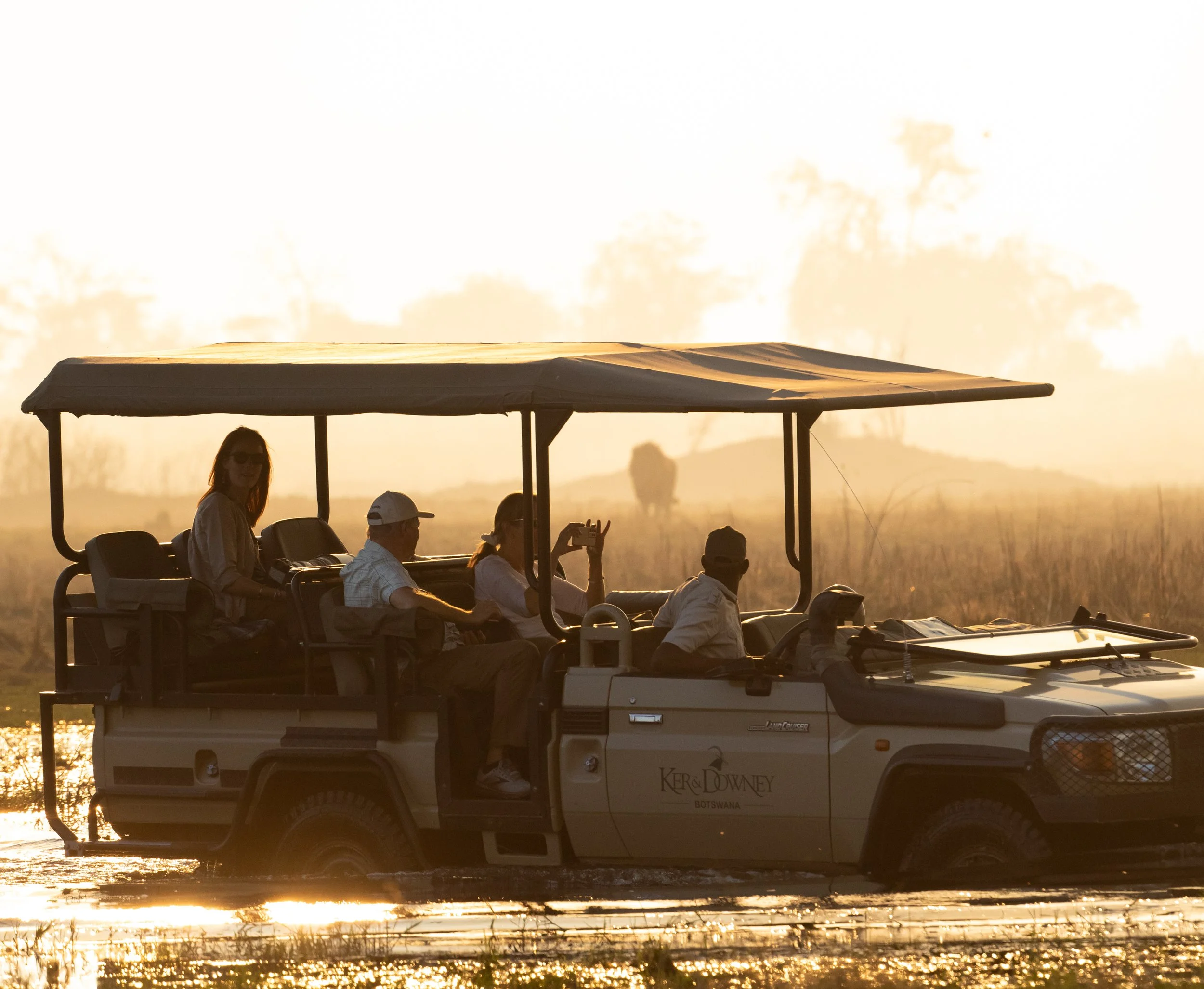 A group of five people riding in an open-air safari vehicle through a river at sunset, with a sign reading 'Ker & Downey Botswana' on the vehicle.