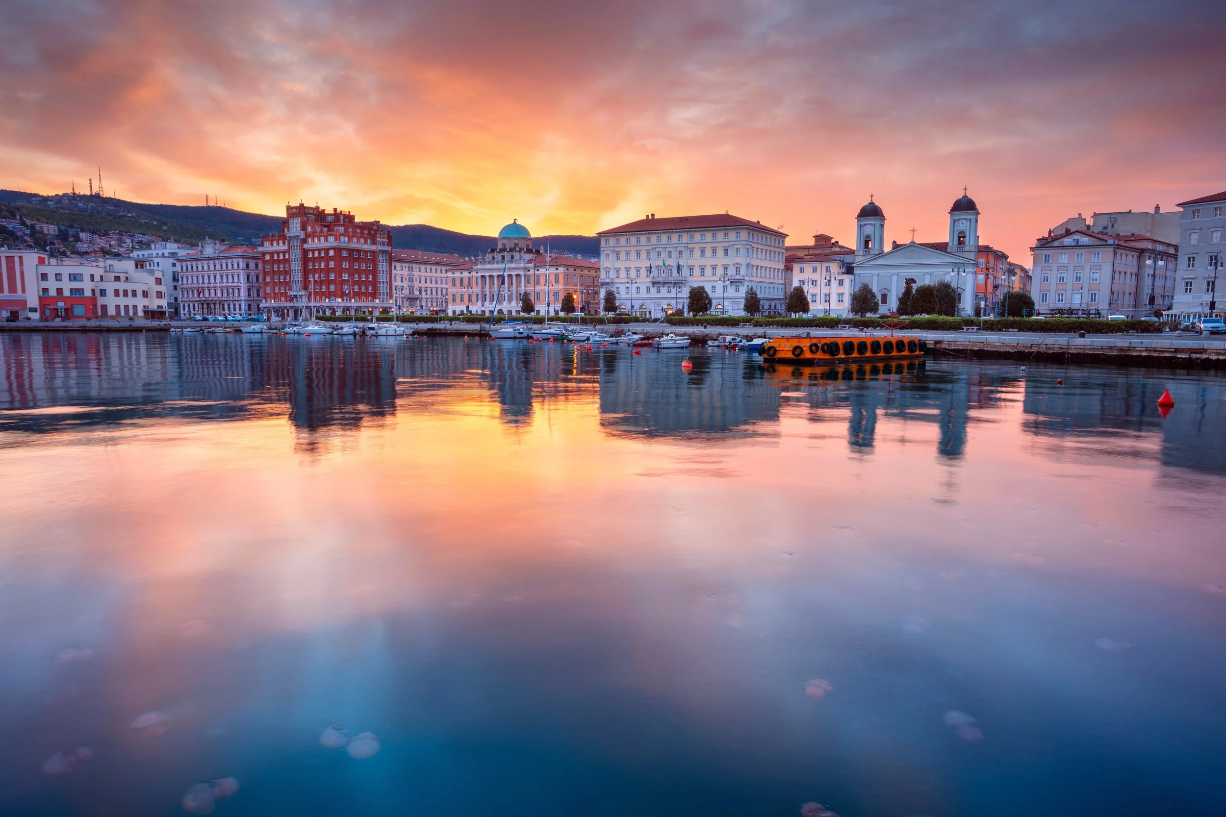 A cityscape with historic buildings and churches along a waterfront at sunset, with reflections on the calm water.