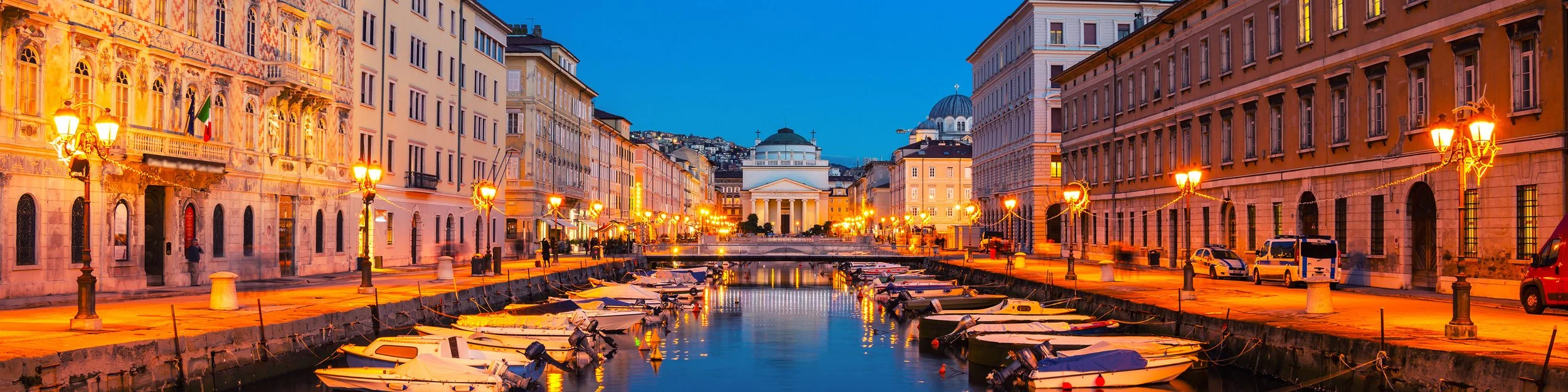 A canal lined with boats docked along both sides, illuminated by street lamps, in a European city during twilight.