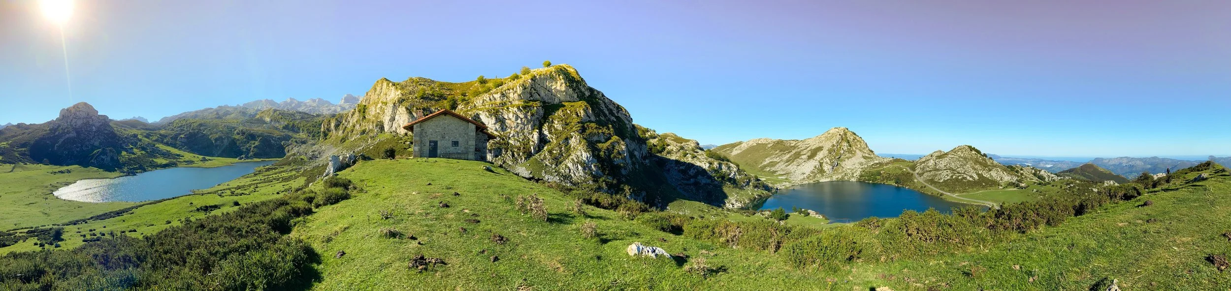 Panoramic view of a mountainous landscape with green hills, rocky peaks, and two lakes, one in the foreground and one in the background, under a clear blue sky.