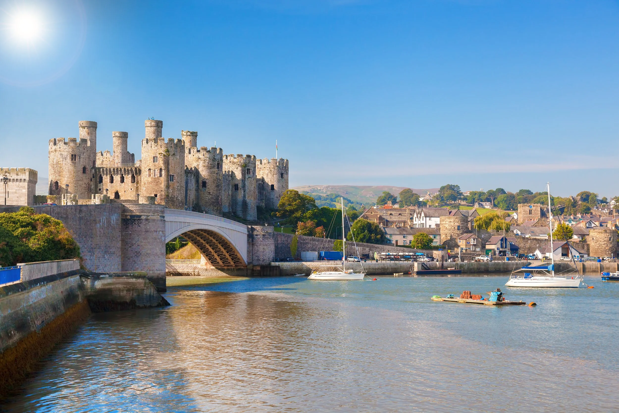 A castle overlooking a river with sailboats and a boat, under a clear blue sky.